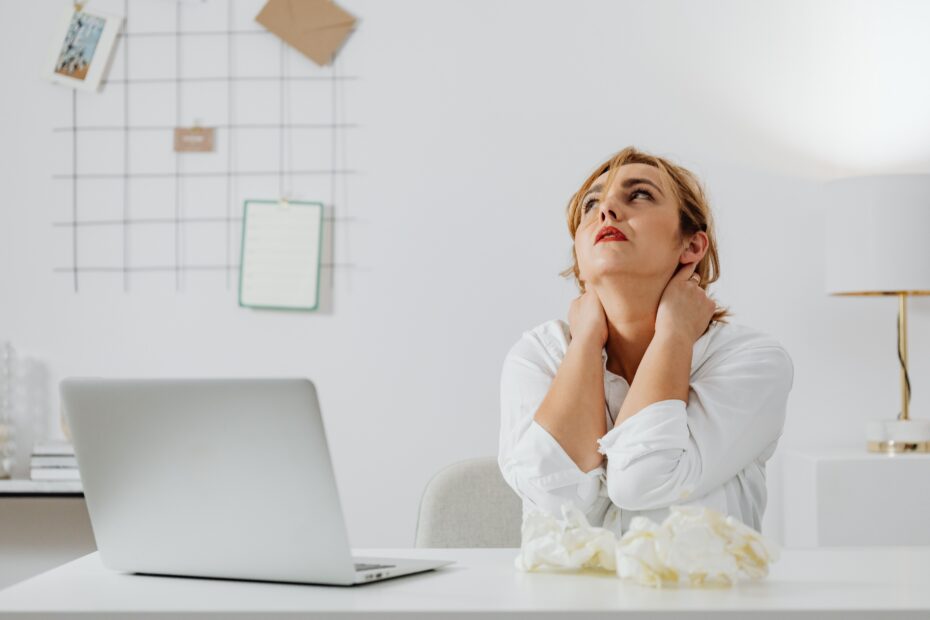 Woman sitting at a desk with a laptop, hand on her neck showing signs of tech neck strain from poor posture during screen work
